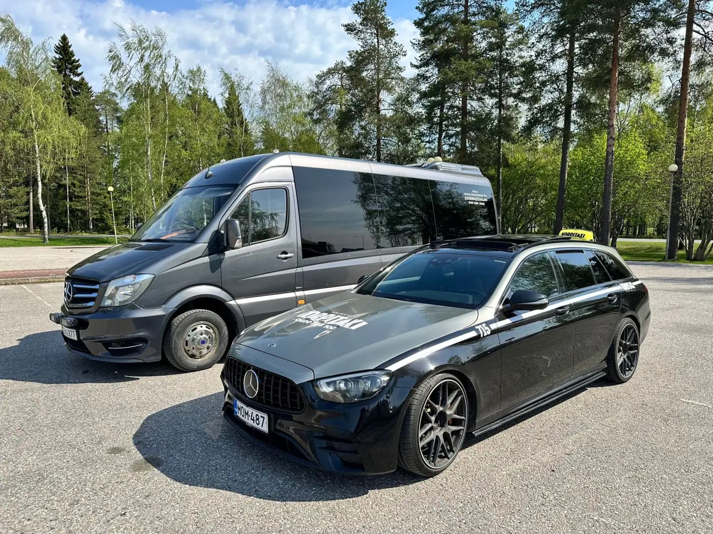 Musta pikkubussi ja taksikyltillä varustettu tumma farmariauto pysäköitynä kesäisen koivikon reunaan